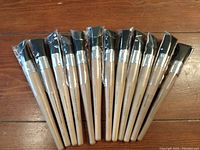 Set of twelve new paint brushes arranged side by side on wooden surface, showing variation in brush shapes and sizes.
