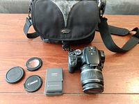 Overview of the Canon EOS Rebel Xti camera with lenses caps, battery charger, and Lowepro camera bag on a wooden table.