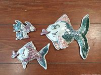Three ceramic fish wall pieces on wooden surface showing varying sizes and colors