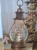 Photo of the 12-inch tall holiday lantern showing the snowman and children figurine scene inside the lantern with bronze-tone metal top and bottom and clear, textured glass.
