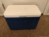 Front and side view of the blue and white Coleman cooler with closed lid on carpet.