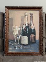 Framed print showing two champagne bottles and two champagne flutes on table surface. Print is behind glass with a decorative carved wooden frame exhibiting signs of wear.