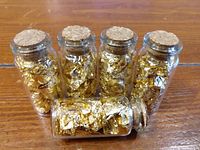 Five small glass vials filled with gold leaf flakes, arranged with one vial lying down in front of the others.