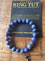 Photo showing a blue lapis lazuli bead bracelet with an attached black ankh charm, displayed on a card referencing King Tut and the healing properties of lapis lazuli.