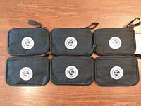 Six black zippered sewing kits with wrist straps and circular emblems, arranged in two rows of three.