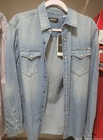 Light blue denim shirt with button front and two chest pockets with button flaps, hanging on a hanger, showing tags from Jack & Jones brand.