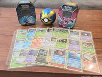 Two Pokémon trading card tins showing characters Dialga and Gyarados, a Poké Ball spherical tin, and 54 Pokémon cards spread out on a wooden surface.