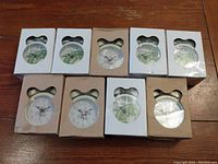 Nine boxed and unboxed white travel clocks with two different styles shown, arranged in rows on wooden surface.