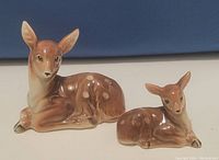 Image of two ceramic deer figurines, a larger doe and a smaller fawn, both resting. Glazed finish, brown with white spots on the fawn.