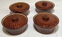 Four McCoy brush brown ceramic ramekins with lids arranged on a white surface, showing round ribbed exterior and textured lids.