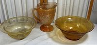 Three amber glass pieces on white cloth background: pitcher with floral etching and two decorative bowls.