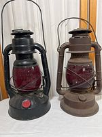 Two vintage Little Wizard oil lamps displayed side by side, one black and one rust-colored, both with red glass globes.