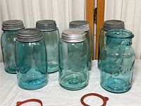 Seven vintage blue Ball Ideal canning jars with metal lids arranged on white cloth, two red rubber sealing rings in front.