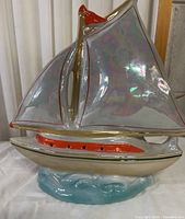 Front view of the iridescent ceramic ship lamp showing shiny reflective sails, gold and red hull details, base shaped like ocean waves.