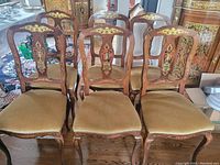 Front and angled front view photo showing the set of six vintage dining chairs with beige upholstered seats and decorative wooden backs with inlay and gilt details.