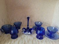 Lot of 8 blue glass items including cups, bowls, plates, bud vase and ashtray/candy dish on beige fabric background.