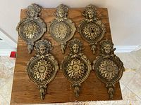 Top view of six solid brass plaques arranged on a wooden board, showing the front side with female heads and floral designs.