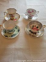 Top-down view showing four bone china teapots and saucers with floral patterns and gilt edges on a white textured cloth.