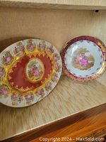 Both the oval and round decorative plates are shown side by side on a wood shelf, highlighting their artwork and gold embellishments