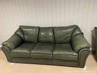 Front view of green leather sofa with three seat cushions and three back cushions against white paneled wall.