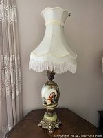 Full view of the Victorian style lamp showing scalloped cream silk shade with fringe and beaded trim, tall hand painted ceramic base, decorative metal fittings, and marble base on table.