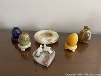 Top view of six polished stone decorative items: three eggs with stands in purple, green and orange, a round bowl/receiver, a heart-shaped trinket dish with carved doves, and a table top lighter with brass top, all on a wooden surface.