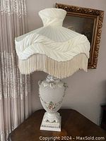 Full view of lamp including ivory pleated fabric shade with fringe trim and white porcelain lamp base with floral and cherub details.