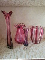 Full view of four ruby cranberry colored hand blown glass vases arranged side by side on fabric background, showing varied shapes and sizes including tall fluted, ruffled lip bouquet, petite trumpet and striped urn vases.