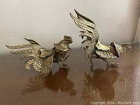 Front and side view of two brass rooster sculptures on wooden surface.