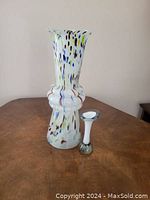 Two art glass vases photographed on wood table against a plain wall: a tall splatter-pattern vase and a small Murano bud vase.