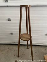 Full view of the wooden tripod plant stand with two tiers, standing on concrete floor against a white garage door. Shows overall shape, dimensions and condition.