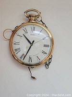 Front view of round brass finish clock with white dial and Roman numerals, chain, and jump ring hanger visible.