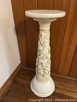 Full view of the white plaster plant pedestal showing the round top, tapering column decorated with grape clusters and vine leaves, and domed base.
