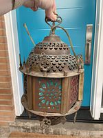 Front-side view of the hexagonal antique brass lamp showing turquoise and red colored glass panels with decorative metal cut-outs and top hanging loop.