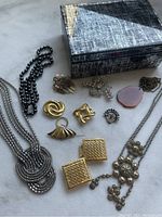 Photo showing assorted silver-tone and gold-tone jewelry pieces including necklaces, earrings, brooches, along with a textured decorative box from Westelm.