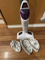 Full view of the white Bissell floor steamer with purple accents standing upright on a wooden floor, accompanied by three mop pads laid out in front.