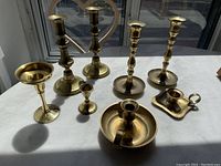 Overall view of all 6 brass candle sticks on a white surface displaying various shapes and sizes.