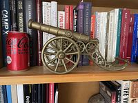 Side view of brass cannon on bookshelf next to a soda can for scale, showing tubular barrel and ornate spoked wheels.