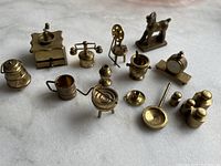 Overall view of 14 brass miniature items arranged on white surface showing various kitchen, weighing, and decorative items.