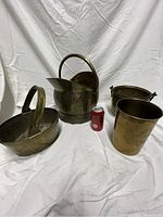 Four brass containers on white cloth, including oval bucket, curved pitcher bucket with backplate, small round bucket, and tall pot. Included Coke can for scale.