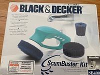 Main boxed Black & Decker Scumbuster Kit showing teal handheld power scrubber and two brush attachments in packaging with logo and info.