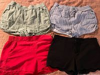 Four pairs of shorts laid flat on a surface: green linen, blue striped linen, red scalloped, black scalloped silk
