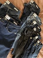 Top view of 10 pairs of Frame jeans arranged on a bed showing label tags and various denim washes. Highlights the different styles from light to dark and black denim.