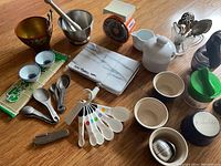Overview of assorted kitchen items including spoons, teapot, citrus juicer, cheese slicer, and scale on table