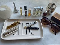 Overview of perfumes, sunglasses, and vanity items including lipsticks and trinkets on white surface with 'SELFIE' dish.