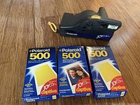 Polaroid JoyCam black instant camera with yellow wrist strap and built-in flash, positioned above three factory-sealed boxes of Polaroid 500 film labeled for JoyCam Captiva use on wooden surface.