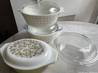 Four vintage Pyrex dishes, two white with green floral design and two clear glass, shown stacked and arranged on a marble surface.