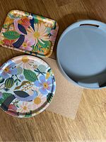 Top view of all three trays on wooden floor: two decorative Rifle Paper Co. trays with floral and bird designs and one grey silicone tray.