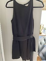 Black sleeveless romper hanging on a wooden hanger, smooth fabric with matching belt tied at waist