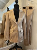 Front view of satin champagne blazer on mannequin with matching pants hanging behind